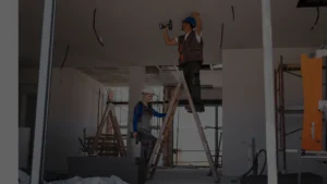 Dois trabalhadores com capacetes e furadeiras utilizando uma escada em uma reforma de uma casa sem riscos estruturais.