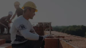 Canteiro de obras pequeno, com trabalhador de capacete amarelo, luvas e tijolos cerâmicos, com segurança de obras.