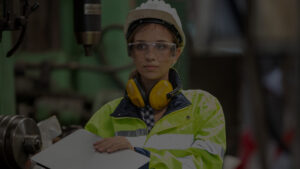 Trabalhadora em ambiente industrial utilizando capacete, óculos de proteção e protetor auricular, realizando inspeção de segurança conforme os requisitos da NR-12